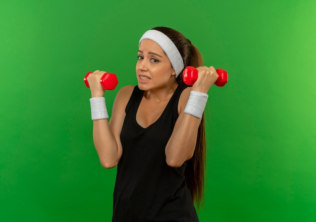 Chica joven fitness en ropa deportiva con diadema sosteniendo dos pesas haciendo ejercicios mirando confundido parado sobre pared verde