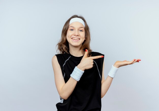 Chica joven fitness en ropa deportiva negra con diadema apuntando con el dedo hacia el lado presentando algo con el brazo de la mano sonriendo de pie sobre la pared blanca