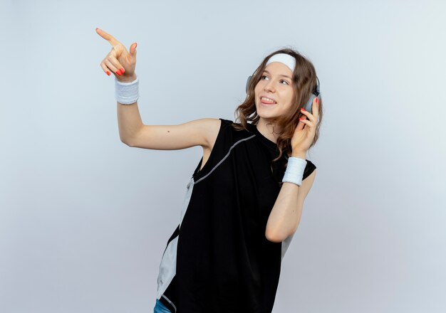 Chica joven fitness en ropa deportiva negra con diadema y auriculares mirando a un lado sonriendo apuntando con el dedo a algo parado sobre la pared blanca