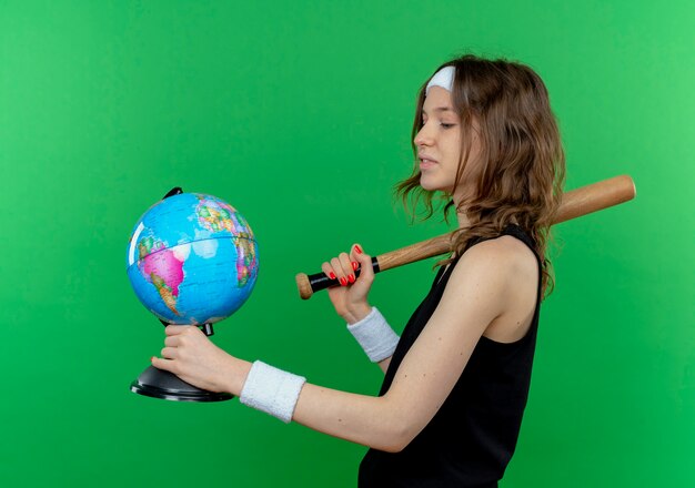 Chica joven fitness en ropa deportiva negra con diadema con bate de béisbol y globo mirándolo con cara seria de pie sobre la pared verde