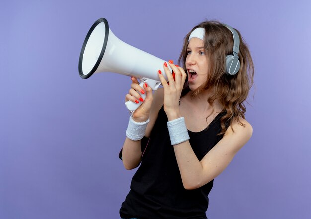 Chica joven fitness en ropa deportiva negra con diadema y brazalete de teléfono inteligente gritando al megáfono de pie sobre la pared azul