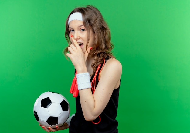 Chica joven fitness en ropa deportiva negra con diadema y cuerda alrededor del cuello sosteniendo un balón de fútbol sonriendo alegremente de pie sobre la pared verde