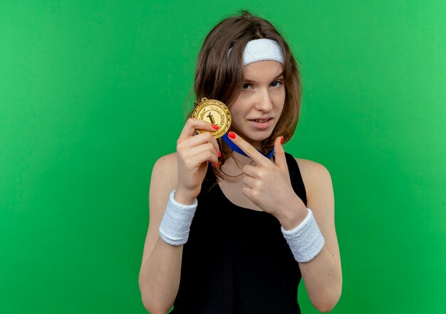 Chica joven fitness en ropa deportiva negra con diadema y medalla de oro alrededor del cuello apuntando con el dedo hacia él mirando confiado de pie sobre la pared verde