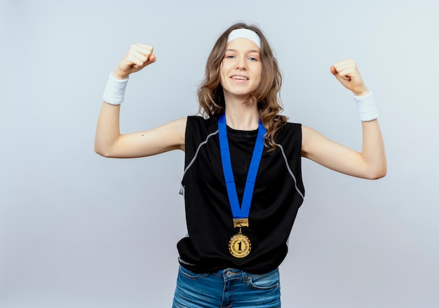 Foto gratuita chica joven fitness en ropa deportiva negra con diadema y medalla de oro alrededor del cuello levantando los puños como un ganador que parece seguro de pie sobre una pared blanca
