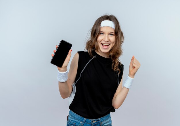 Chica joven fitness en ropa deportiva negra con diadema mostrando smartphone apretando el puño feliz y emocionado de pie sobre la pared blanca