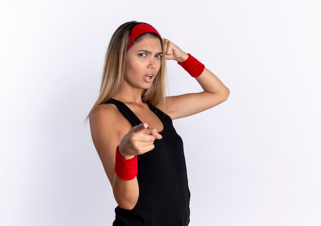 Chica joven fitness en ropa deportiva negra y diadema roja apuntando con el dedo índice mirando con cara seria de pie sobre una pared blanca