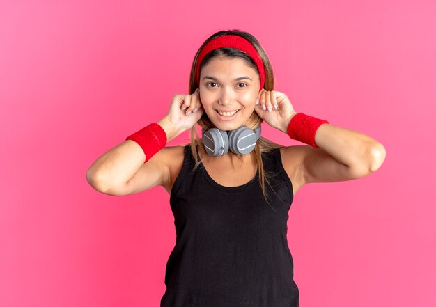 Chica joven fitness en ropa deportiva negra y diadema roja con auriculares divirtiéndose sonriendo de pie sobre la pared rosa