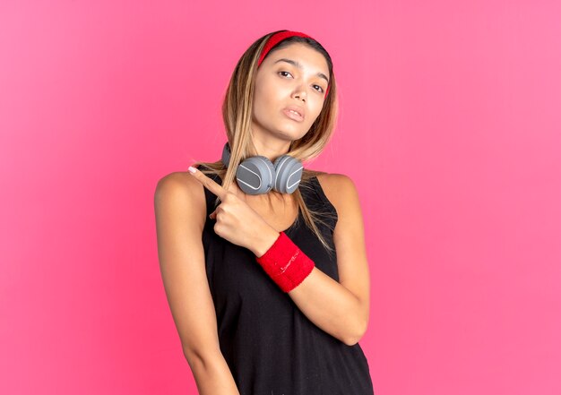 Chica joven fitness en ropa deportiva negra y diadema roja con auriculares mirando confiado apuntando hacia atrás sobre rosa