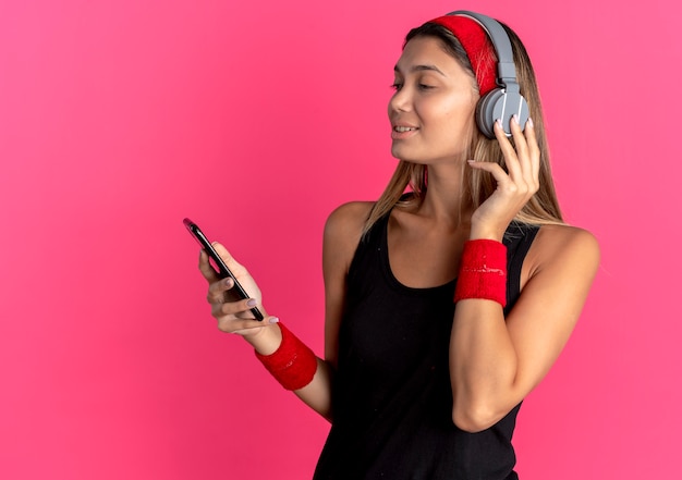 Chica joven fitness en ropa deportiva negra y diadema roja con auriculares mirando la pantalla de su teléfono inteligente buscando música sobre rosa