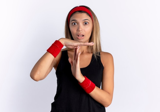 Chica joven fitness en ropa deportiva negra y diadema roja confundida haciendo gesto de tiempo de espera con las manos de pie sobre la pared blanca