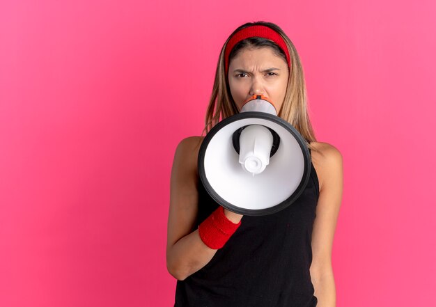 Chica joven fitness en ropa deportiva negra y diadema roja gritando al megáfono parado sobre la pared rosa