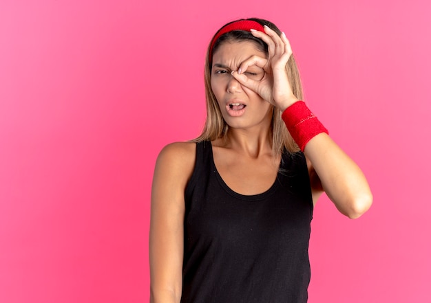 Chica joven fitness en ropa deportiva negra y diadema roja haciendo el signo de ok mirando a la cámara a través de este cantar con expresión confusa de pie sobre la pared rosa