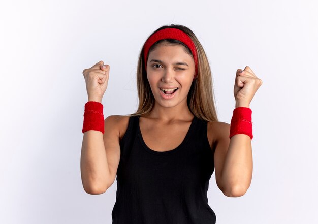 Chica joven fitness en ropa deportiva negra y diadema roja lookign apretando los puños guiñando un ojo y sonriendo de pie sobre la pared blanca