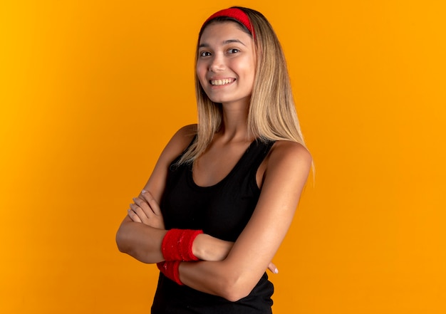 Chica joven fitness en ropa deportiva negra y diadema roja mirando confiado sonriendo con los brazos cruzados de pie sobre la pared naranja