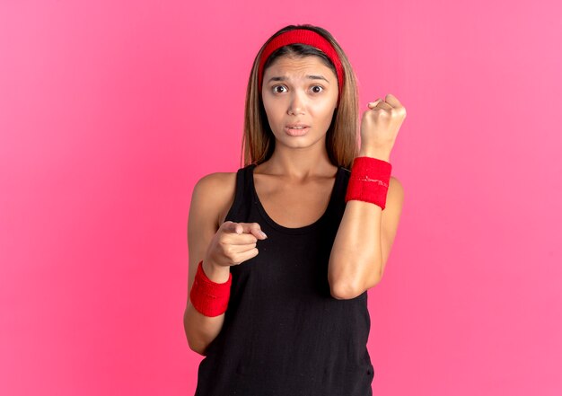 Chica joven fitness en ropa deportiva negra y diadema roja mirando confundido apretando el puño apuntando con el dedo a la cámara sobre rosa