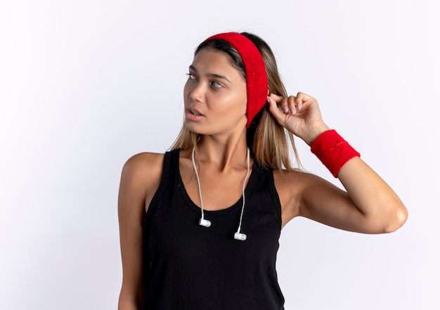 Chica joven fitness en ropa deportiva negra y diadema roja mirando a un lado con cara seria de pie sobre la pared blanca