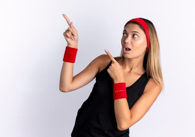 Chica joven fitness en ropa deportiva negra y diadema roja mirando sorprendido apuntando con el dedo índice a algo parado sobre la pared blanca