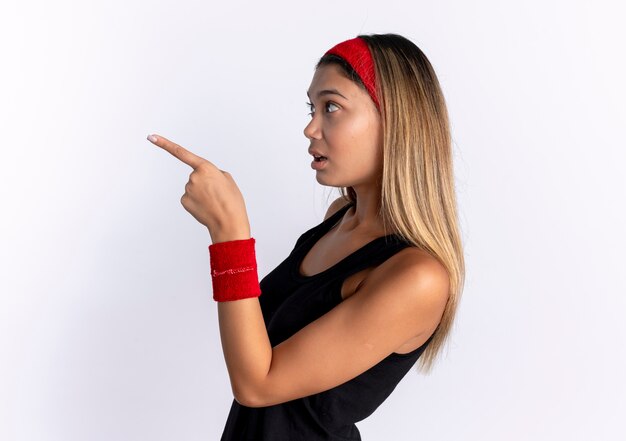 Chica joven fitness en ropa deportiva negra y diadema roja de pie hacia los lados apuntando con el dedo a algo sorprendido sobre la pared blanca