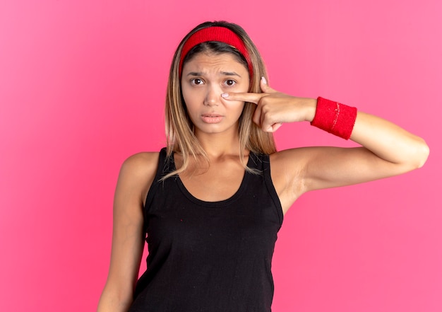 Chica joven fitness en ropa deportiva negra y diadema roja pointign con el dedo en la nariz con expresión triste de pie sobre la pared rosa