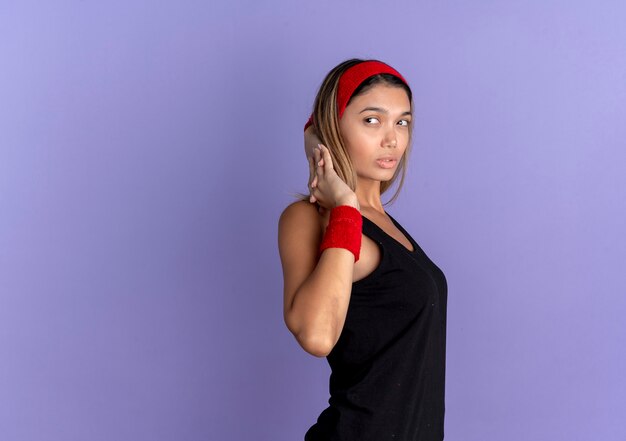 Chica joven fitness en ropa deportiva negra y diadema roja que parece segura de estirarse de pie sobre la pared azul