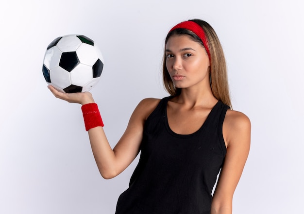 Chica joven fitness en ropa deportiva negra y diadema roja sosteniendo un balón de fútbol con cara seria de pie sobre una pared blanca