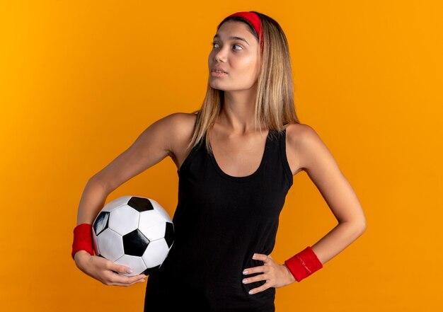 Chica joven fitness en ropa deportiva negra y diadema roja sosteniendo un balón de fútbol mirando a un lado con expresión segura sobre naranja