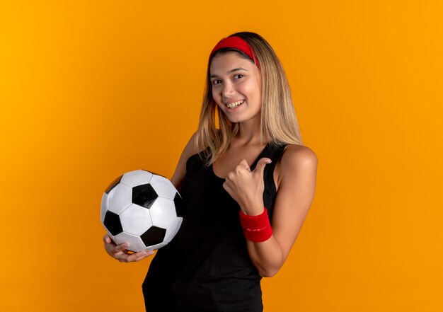 Chica joven fitness en ropa deportiva negra y diadema roja sosteniendo un balón de fútbol sonriendo mostrando los pulgares para arriba de pie sobre la pared naranja