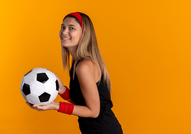 Chica joven fitness en ropa deportiva negra y diadema roja sosteniendo un balón de fútbol con una sonrisa en la cara de pie sobre la pared naranja