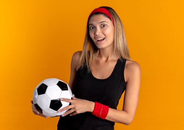 Chica joven fitness en ropa deportiva negra y diadema roja sosteniendo un balón de fútbol sorprendido con cara feliz sobre naranja