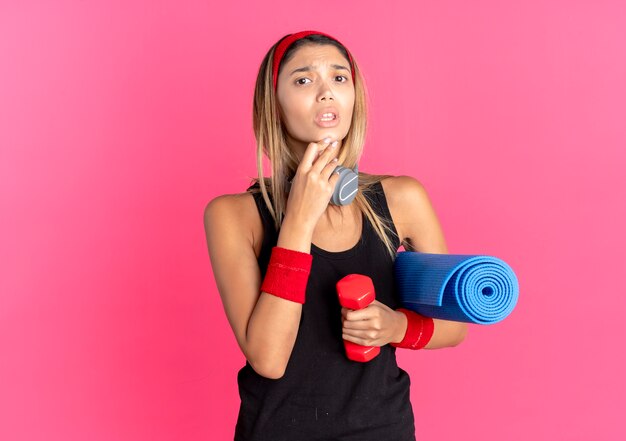 Chica joven fitness en ropa deportiva negra y diadema roja sosteniendo mancuernas y estera de yoga confundida sobre rosa