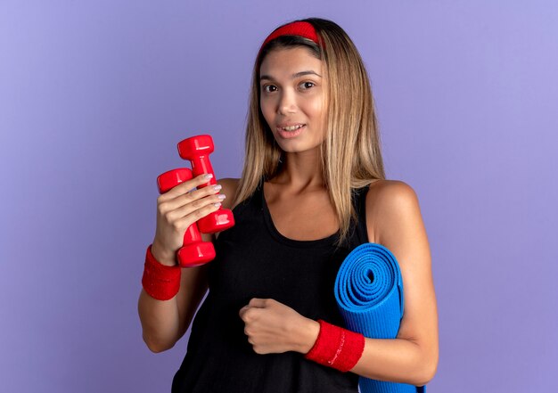 Chica joven fitness en ropa deportiva negra y diadema roja sosteniendo pesas y estera de yoga mirando a la cámara sonriendo confiado de pie sobre la pared azul