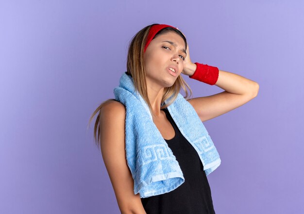 Chica joven fitness en ropa deportiva negra y diadema roja con una toalla alrededor del cuello que parece confundida con la mano en la cabeza por error de pie sobre la pared azul