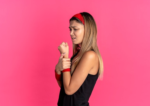 Chica joven fitness en ropa deportiva negra y diadema roja tocando su muñeca sintiendo dolor sobre rosa