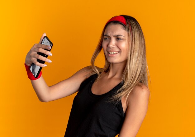 Chica joven fitness en ropa deportiva negra y diadema roja tomando selfie con smartphone mirando disgustado parado sobre pared naranja