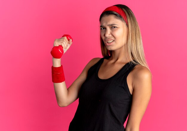 Chica joven fitness en ropa deportiva negra y diadema roja trabajando con mancuernas mirando confundido parado sobre pared rosa