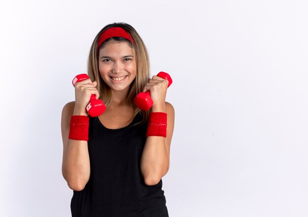 Chica joven fitness en ropa deportiva negra y diadema roja trabajando con pesas lookign sonriendo alegremente de pie sobre la pared blanca