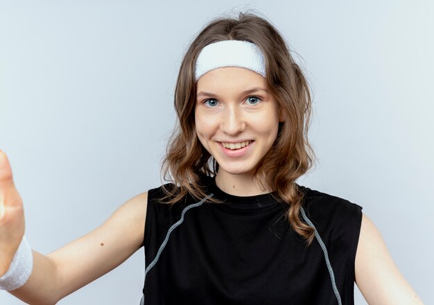Chica joven fitness en ropa deportiva negra con diadema con rostro serio con los brazos cruzados sobre la pared blanca
