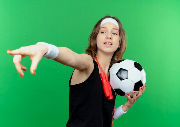 Chica joven fitness en ropa deportiva negra con diadema y saltar la cuerda alrededor del cuello sosteniendo un balón de fútbol mirando algo apuntando con el dedo parado sobre la pared verde