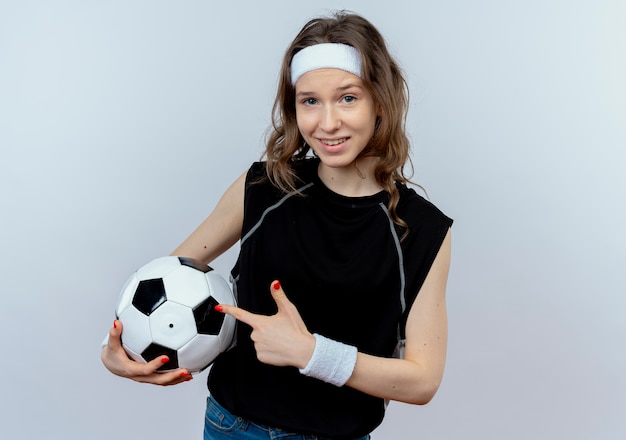 Chica joven fitness en ropa deportiva negra con diadema sosteniendo un balón de fútbol apuntando con el dedo sonriendo alegremente de pie sobre la pared blanca