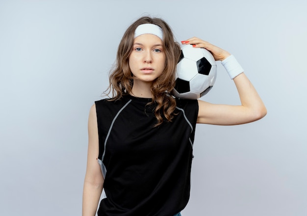 Chica joven fitness en ropa deportiva negra con diadema sosteniendo un balón de fútbol con cara seria de pie sobre la pared blanca