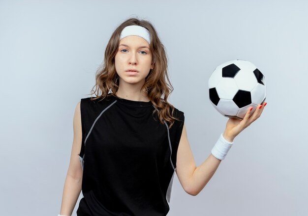 Chica joven fitness en ropa deportiva negra con diadema sosteniendo un balón de fútbol con cara seria de pie sobre la pared blanca