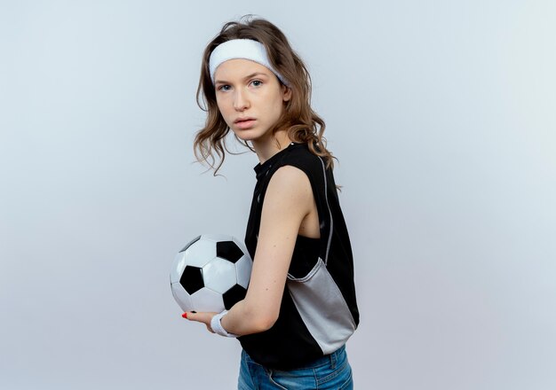 Chica joven fitness en ropa deportiva negra con diadema sosteniendo un balón de fútbol con cara seria de pie sobre la pared blanca