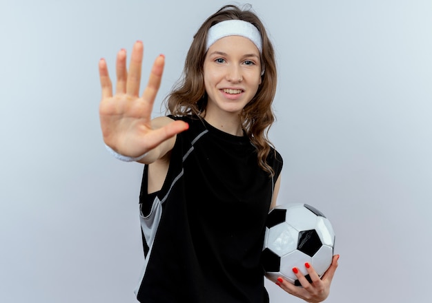 Chica joven fitness en ropa deportiva negra con diadema sosteniendo un balón de fútbol haciendo señal de stop con la mano abierta sonriendo de pie sobre la pared blanca