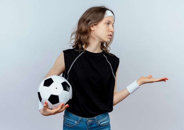 Chica joven fitness en ropa deportiva negra con diadema sosteniendo un balón de fútbol mirando a un lado con el brazo extendido mientras pregunta de pie sobre la pared blanca