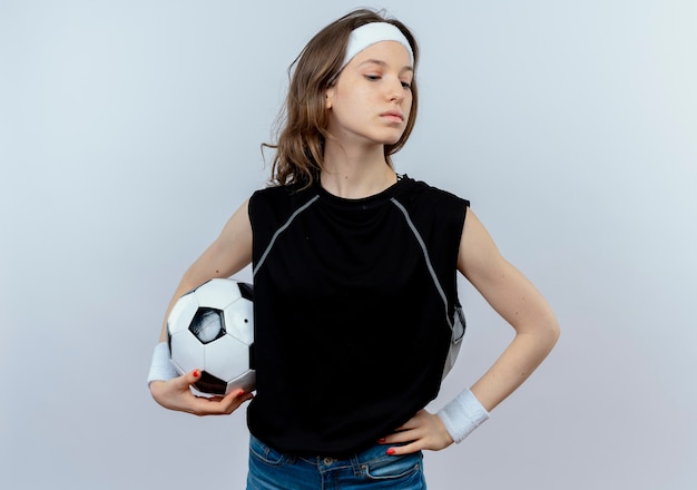 Chica joven fitness en ropa deportiva negra con diadema sosteniendo un balón de fútbol mirando a un lado seguro de pie sobre la pared blanca