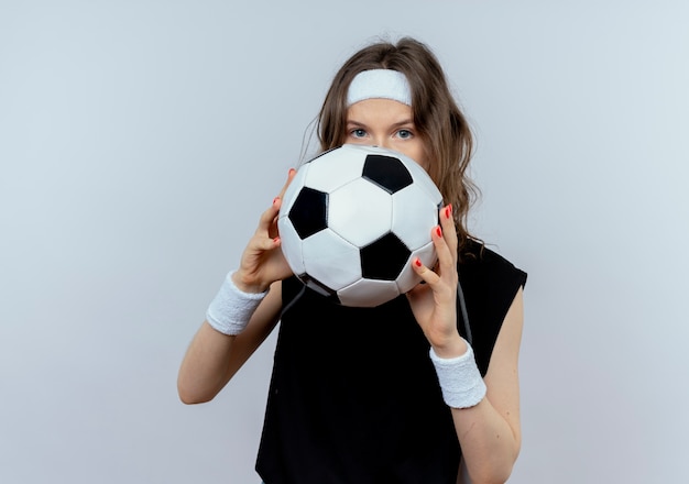 Chica joven fitness en ropa deportiva negra con diadema sosteniendo un balón de fútbol ocultando su rostro parado sobre una pared blanca