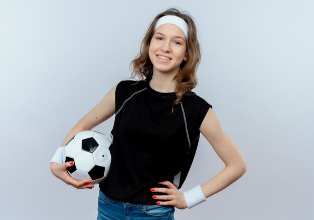 Chica joven fitness en ropa deportiva negra con diadema sosteniendo un balón de fútbol sonriendo confiado de pie sobre la pared blanca