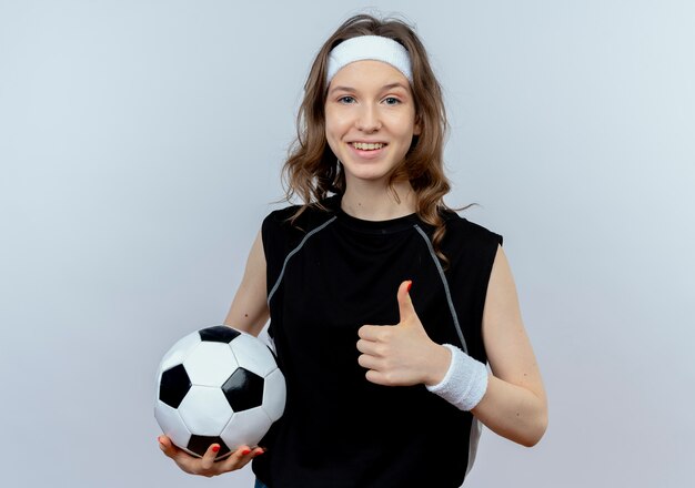 Chica joven fitness en ropa deportiva negra con diadema sosteniendo un balón de fútbol sonriendo mostrando los pulgares para arriba de pie sobre la pared blanca