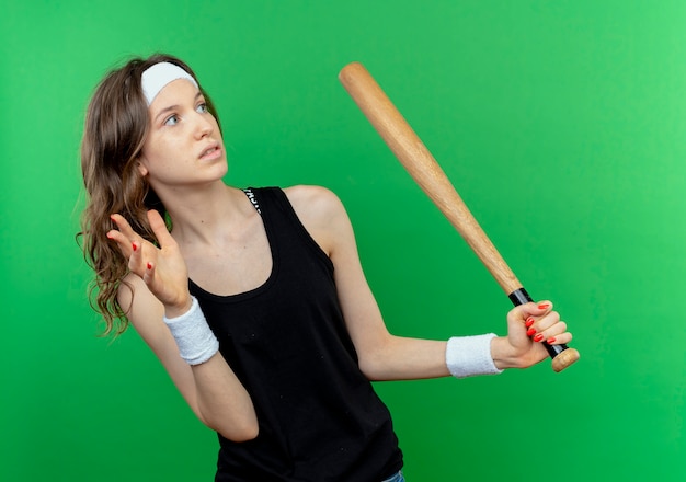 Foto gratuita chica joven fitness en ropa deportiva negra con diadema sosteniendo bate basaball mirando a un lado asustado de pie sobre la pared verde