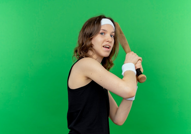 Foto gratuito chica joven fitness en ropa deportiva negra con diadema sosteniendo un bate de béisbol confundido de pie sobre la pared verde